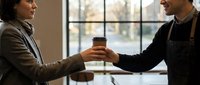 Young male barista of South Asian descent in black apron handing takeaway coffee cup to male customer of Middle Eastern descent in brown jacket indoors near window.