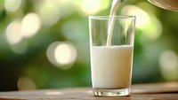 Fresh milk being poured into clear glass on wooden surface with natural green bokeh background for healthy breakfast and dairy nutrition concepts.