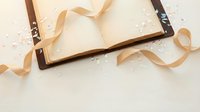 Open vintage book with flowing golden ribbons on white surface with scattered confetti for romantic storytelling and celebration themes.