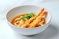 Tempura shrimp udon noodle soup in white ceramic bowl with clear broth, garnished with fresh herbs and black pepper on light background.