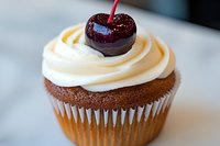 Chocolate cupcake topped with swirled vanilla buttercream frosting and garnished with glazed dark cherry, close-up view on white surface showing texture and detail.