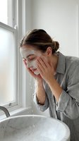 Woman with brown hair in casual gray shirt washing face with water over white sink in bright bathroom with window light, skincare routine.