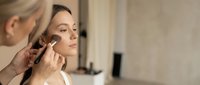 Professional makeup artist applying foundation with brush to young Caucasian woman face in beauty salon, skincare routine.