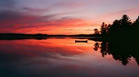 Serene lake at sunset with vibrant orange and red sky reflecting on calm water, silhouetted evergreen trees and moored boat creating peaceful nature scene.