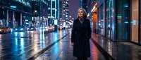 Young blonde woman in black coat walking on wet city street at night with neon lights and urban architecture creating atmospheric downtown scene.
