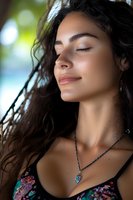 Young woman with long dark wavy hair and Latin ethnicity wearing jeweled pendant necklace outdoors with blurred green foliage background, eyes closed in peaceful expression for wellness and lifestyle content.