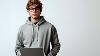 Young male professional in gray hoodie and glasses working on laptop against white background, modern minimalist workspace setting for remote work and technology concepts.