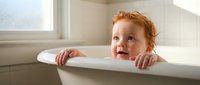 Adorable redhead toddler with curly hair peeking over white bathtub edge during bath time in bright modern bathroom with natural window light.