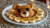 Playful bear-shaped pancake breakfast decorated with chocolate chips, banana slices on white plate. Creative food styling for children's meal on granite counter.