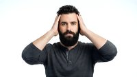 Caucasian male with full dark beard and black hair, wearing dark gray henley shirt, hands on head in thoughtful or stressed pose against white background for portrait and lifestyle use.