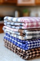 Stack of folded kitchen towels in various plaid patterns including blue, red, and brown on white backgrounds, arranged neatly on countertop.