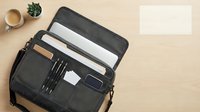 Professional gray laptop bag open displaying organized compartments with laptop, documents, and business accessories on wooden desk with coffee and plant.