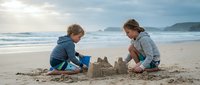 Two young children building sandcastles together on scenic ocean beach during summer vacation with waves and mountains in background.