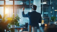 Businessman in dark suit writing on transparent board in modern office with plants and sunset light through windows.
