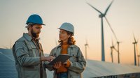 Engineers discussing project on digital tablet at renewable energy site with solar panels and wind turbines during sunset, showcasing sustainable technology development.
