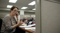 Professional businesswoman talking on phone while working at office desk with documents and computer in modern workplace environment.