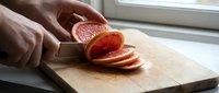 Hands slicing fresh grapefruit on wooden cutting board near window, food preparation and healthy eating concept.