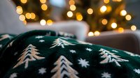 Cozy winter atmosphere with dark green knitted blanket featuring white Christmas tree and snowflake pattern against warm glowing bokeh lights background.