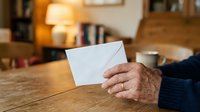 Close-up of mature hands holding blank white envelope on wooden table indoors, suitable for mail, correspondence, greeting card, invitation, and business communication concepts.