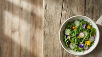 Edible flowers and fresh greens in white bowl on rustic wooden table, healthy salad with radish slices and colorful blooms for culinary and wellness content.