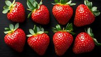 Fresh ripe strawberries with green leaves arranged in rows on dark wooden background, macro food photography showing vibrant red color and texture.