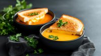 Creamy pumpkin soup served in black bowls with grilled cheese sandwich and fresh parsley garnish on dark background, cozy autumn comfort food.