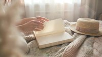 Woman's hands turning pages of open book on wicker tray with straw hat in soft natural sunlight creating peaceful reading moment for relaxation and leisure.
