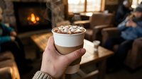 Hand holding hot chocolate with marshmallows by cozy fireplace in warm living room during winter evening creating comfort and relaxation atmosphere.