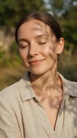 Middle-aged woman with brown hair wearing beige linen shirt, eyes closed with peaceful expression outdoors in natural sunlight with blurred green foliage background.