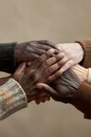 Close up of diverse hands joined together showing unity and support, wearing warm knit sweaters against neutral background, representing community and connection.