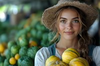 Young woman in straw hat holding fresh yellow pumpkins at farmers market, with green produce display in background. Natural lighting highlights genuine smile.
