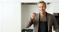 Man in apron tasting food with wooden spoon in modern kitchen, professional chef or cooking instructor demonstrating culinary technique.