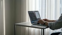 Professional businessman typing on laptop computer at modern office desk near window with natural lighting for remote work and business productivity.