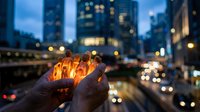 Hands holding glowing fire against blurred city skyline at dusk, suitable for concepts of energy, hope, innovation, and urban themes.