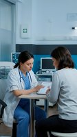 Female doctor in white coat consulting with patient using tablet in modern hospital room discussing medical treatment and healthcare options.