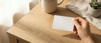 Hand holding blank white card on wooden desk with coffee mug and plant creating minimalist workspace scene for business branding and design mockups.
