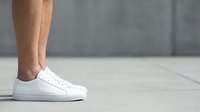 Male legs in white leather sneakers standing on light wooden floor against gray concrete wall background for fashion and footwear advertising.