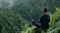 Young Asian woman meditating in lotus position on mountain cliff edge overlooking green forest valley, wearing casual sportswear, practicing mindfulness and yoga outdoors.