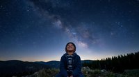 Man in dark jacket gazing upward at starry night sky with milky way galaxy visible above forested mountains, suitable for astronomy, inspiration, and wonder concepts.
