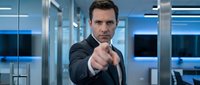 Confident businessman in dark suit pointing finger at camera in modern office with blue lighting and glass walls for corporate presentations.