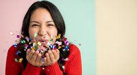 Young Asian woman in red sweater blowing colorful confetti from hands against pastel background celebrating joy and happiness for party events.