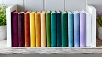 Colorful hardcover books arranged in rainbow spectrum order on white wooden shelf, creating gradient from purple through yellow to blue tones. Minimalist library decor.