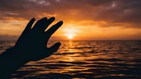 Hand silhouette reaching toward golden sunset over ocean waves, dramatic sky with warm colors, peaceful moment of connection with nature at beach.