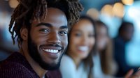 Young African American man with dreadlocks smiling confidently in modern office environment with diverse team of colleagues in background for business concepts.