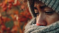 Close-up portrait of African American woman wrapped in warm knitted scarf and hat against autumn foliage background with soft bokeh effect.