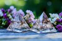 Crystal pyramids with natural inclusions on wooden surface surrounded by purple and pink flowers against blurred green garden background.