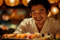 Asian male chef smiling warmly while presenting freshly prepared sushi rolls on wooden board, with soft bokeh lights creating intimate restaurant atmosphere.