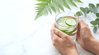 Hands holding refreshing cucumber water glass with green plants on white background for healthy lifestyle and wellness concepts.