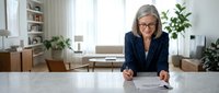 Professional mature businesswoman with glasses reviewing contract documents at modern office desk in bright workspace with plants.