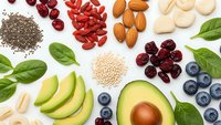 Healthy superfood ingredients including avocado, nuts, seeds, berries and leafy greens arranged on white background for nutritious meal planning.